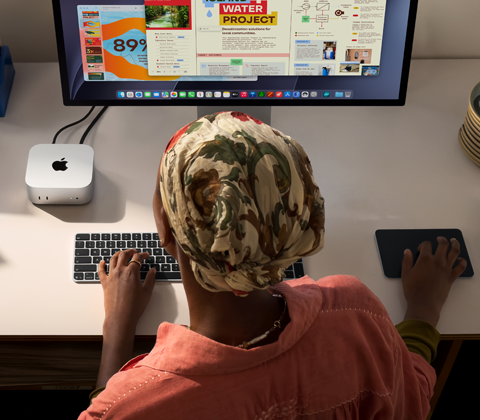 A person typing at a desk with a Mac mini, Studio Display connected, Magic Keyboard, Magic Trackpad, various items