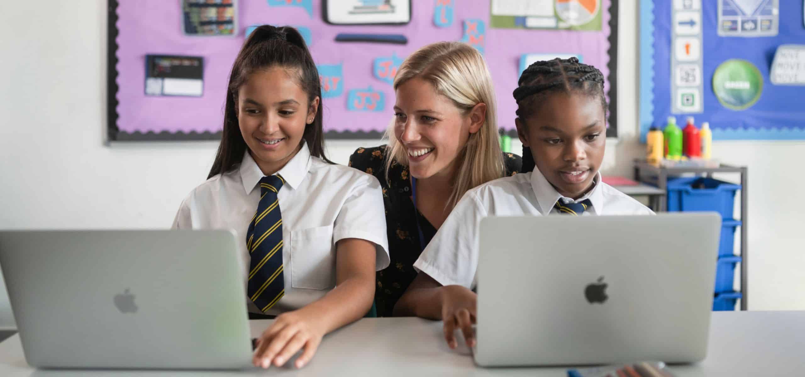 Happy teacher supporting pupils with their work whilst they use their Mac devices in the classroom.