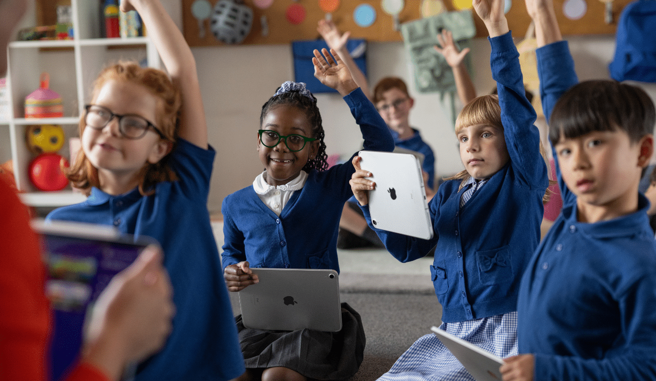 Classroom of children with raised hands, working on iPad
